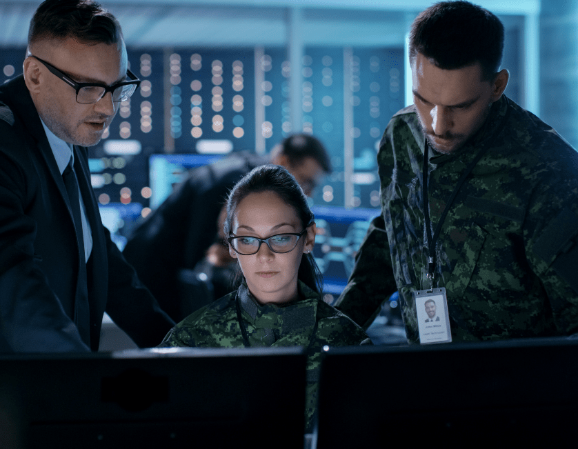Girl in combat suit sitting at computer with two men behind working with her